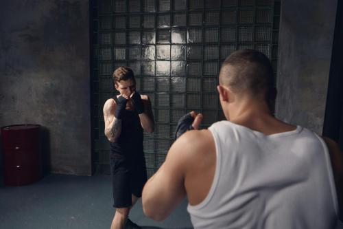 Back view of unrecognizable male fighter with broad muscular shoulders fighting against young Caucasian guy with tattoos wearing black outfit and bandages. Sports, martial arts and competition